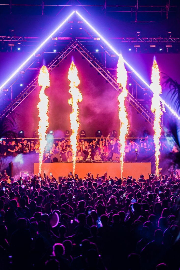 Escenario triangular iluminado en violeta con cuatro columnas de fuego.