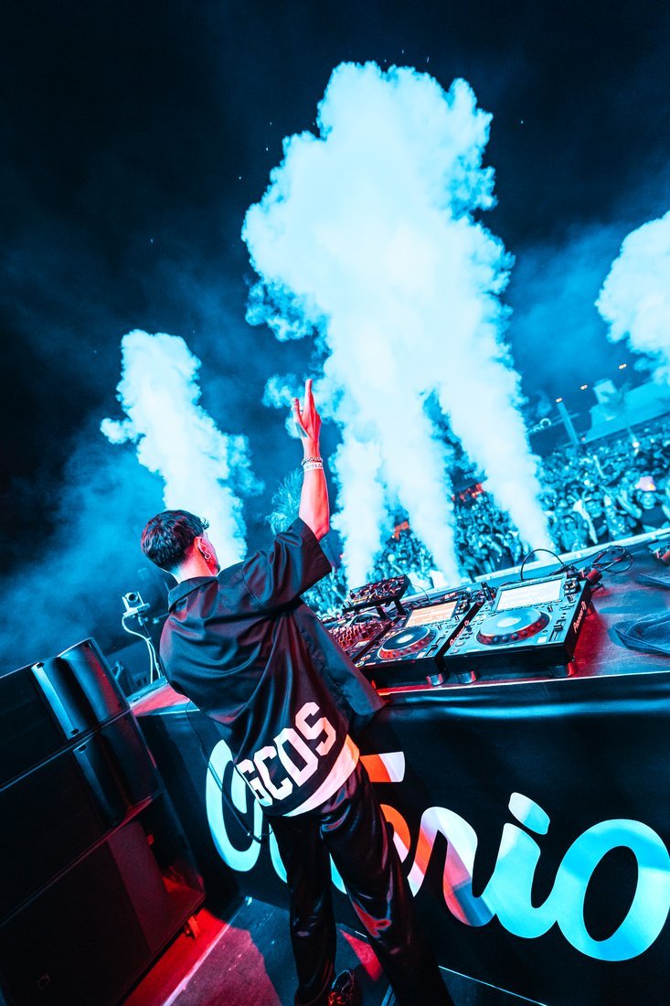 Desde la cabina, un DJ levanta el brazo frente al público y al humo azul.